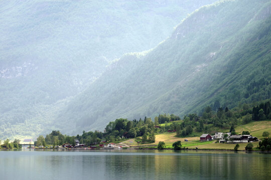 Sunbeams illuminate a peaceful fjord village in Norway, where lush mountains, calm water, and scattered farms create a serene Scandinavian landscape.