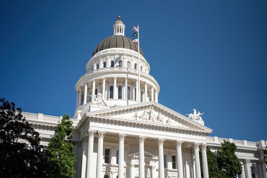 The scene shows the California State Capitol in Sacramento under a clear sky with the flag flying and sunlight shining on the building