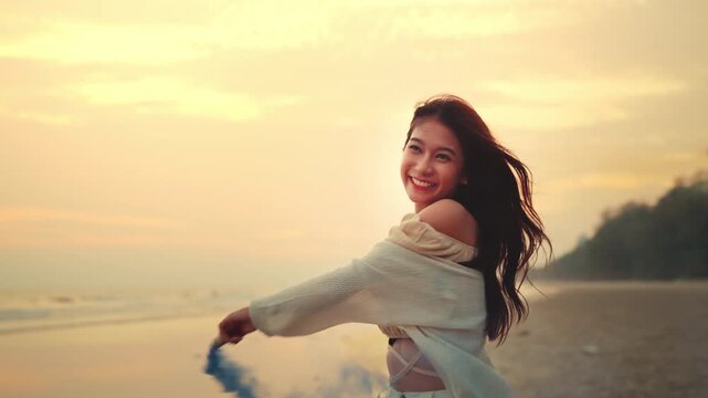 Happy Asian woman holding and playing with colorful smoke bombs on the beach during sunset. Young female enjoying summer vacation with blue and orange smoke flares. Creative celebration and freedom