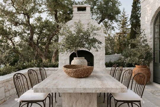 Outdoor dining terrace featuring a limestone manor house with a large stone table, decorative bowl, and wrought iron chairs surrounded by lush greenery