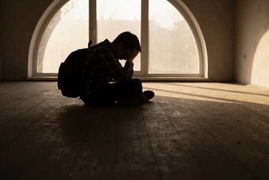 Silhouette of person with backpack sitting in empty room. Sad man with head in hands in front of window. Mental health and loneliness concept