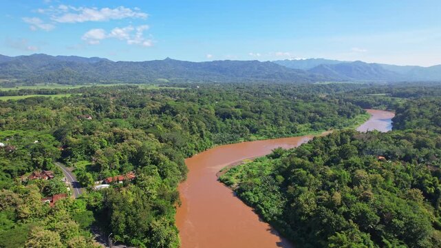 Aerial drone footage of brown color Progo river, in Sleman regency, Yogyakarta area, Java island, Indonesia, with wide green forest, few houses and a road, Kulon Progo mountains in the background