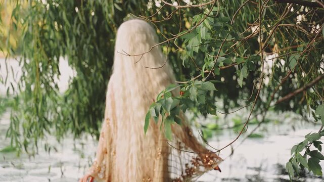 fantasy woman river nymph walking in swamp water view through green foliage tree branches. mystery fairy elf girl in wild nature tree branch leaves. back view wet beige golden dress long blonde hair.