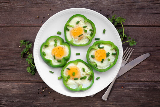 Healthy breakfast concept with eggs in green peppers on a plate. Above view table scene over a dark wood background.