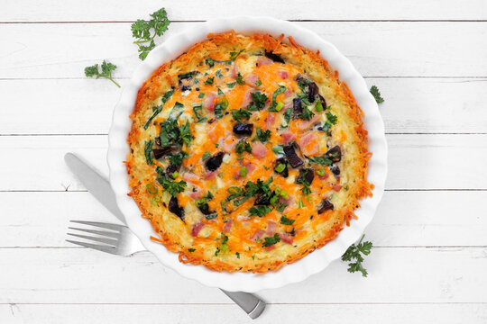 Breakfast quiche with ham, mushrooms, and onions made in a hashbrown shell. Top down view table scene on a white wood background.