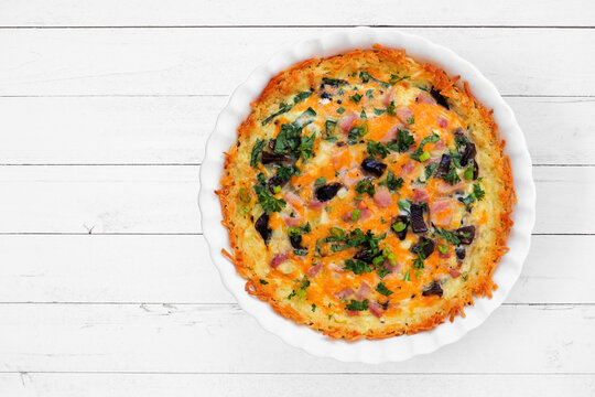 Breakfast quiche with ham, mushrooms, and onions made in a hashbrown shell. Overhead view on a white wood background.