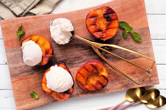 Sweet grilled peaches with vanilla ice cream on a wooden platter. Top down view table scene on a white wood background.