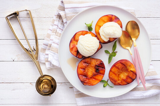 Sweet grilled peaches with vanilla ice cream. Above view table scene on a white wood background.