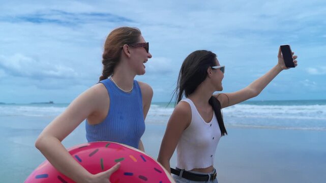 Two young female friends wearing sunglasses taking smartphone selfie and holding colorful inflatable donut pool float at beautiful sandy beach during sunny summer vacation holiday trip.