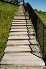 Obraz premium Sunlit wooden stairs ascending grassy hill, close perspective emphasizing planks and black railing, ideal route for hikers and nature walkers seeking viewpoint and quiet escape