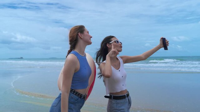 Two young female friends wearing sunglasses taking smartphone selfie and holding colorful inflatable donut pool float at beautiful sandy beach during sunny summer vacation holiday trip.