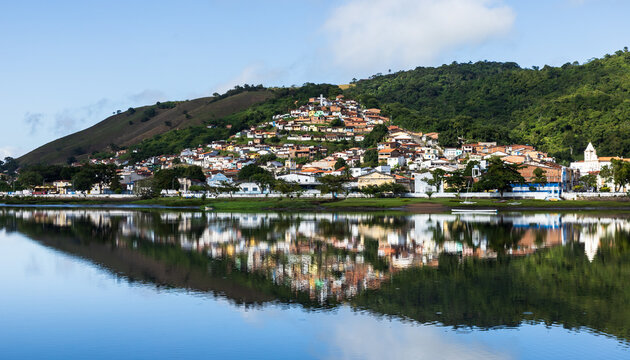 Scenic view of a colorful hillside town reflected in calm river water, surrounded by lush green vegetation, forested hills, traditional houses, church towers and a bright blue sky with soft clouds.