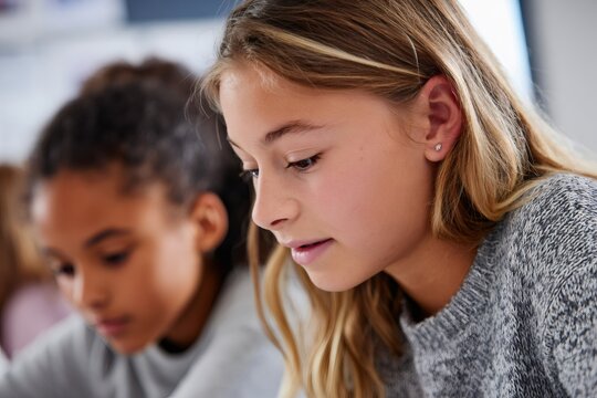 Close up of a focused young schoolgirl studying in a classroom. Elementary student taking a test or writing with a classmate in the background. Education and learning concept