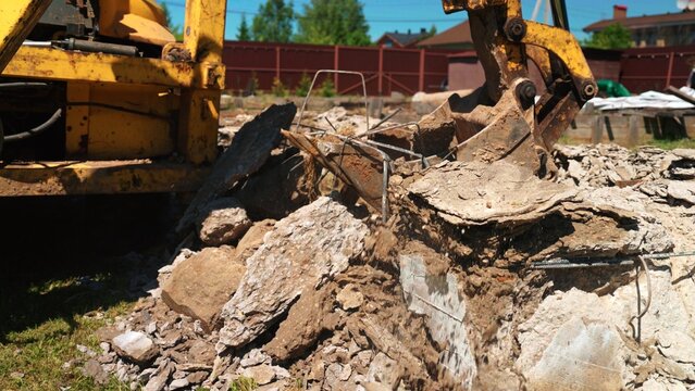 Excavator bucket digging into a pile of broken concrete, rebar, and rubble during demolition work on a construction site under a clear blue sky, showing urban renewal activity
