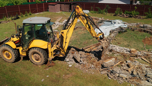 Yellow backhoe excavator performing demolition work in a residential backyard, digging and moving concrete debris and soil during construction site preparation on a sunny day