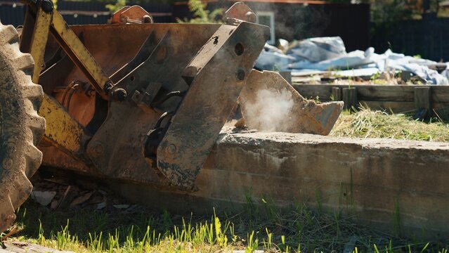 Close-up of a backhoe loader bucket pressing against a reinforced concrete foundation with dust rising during residential demolition, showing heavy machinery, structural removal and site preparation.