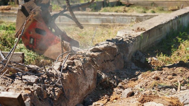 Heavy machinery with an industrial hydraulic breaker demolishing a concrete foundation and reinforced rebar structure at a construction site, creating rubble and debris