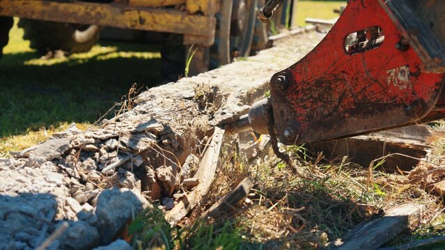 Hydraulic breaker breaking a reinforced concrete foundation beam during residential demolition, with exposed rebar, concrete rubble and heavy construction equipment.