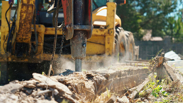 Close-up of a hydraulic demolition attachment breaking an old concrete foundation, with dust, rubble and exposed reinforcement at an outdoor worksite.