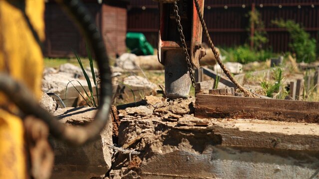 Hydraulic breaker tool breaking up concrete foundation, creating dust and debris during demolition work at a construction site in bright daylight