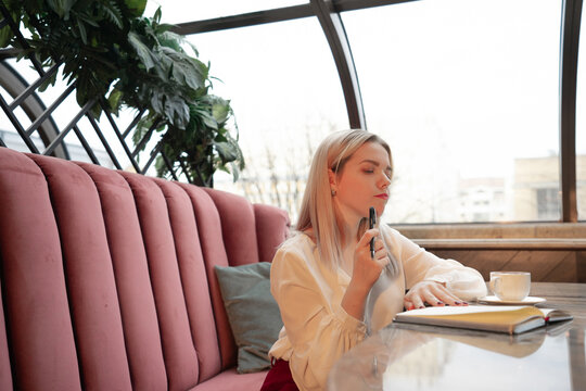 Dreamy young woman writing down future plans and goals in own diary resting in cozy cafeteria