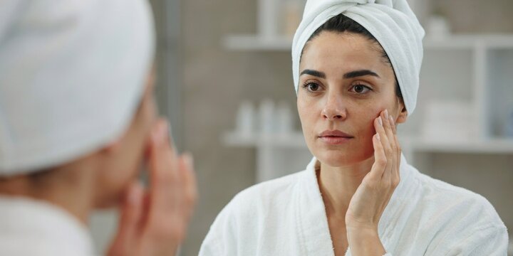 Reflection of young woman looking in bathroom mirror touching face with hands after showering.