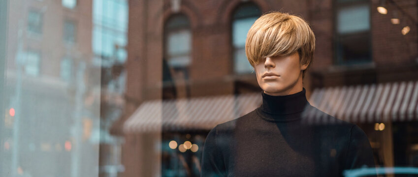 Mannequin behind shop window reflection of a city street with warm brown buildings, featuring a male dummy in a black turtleneck for fashion retail display and boutique advertising