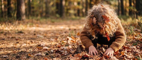 Obraz premium Child playing in autumn leaves during a warm sunny day in the forest, wearing a cozy brown knitted sweater with foliage in curly hair, perfect for seasonal family lifestyle designs