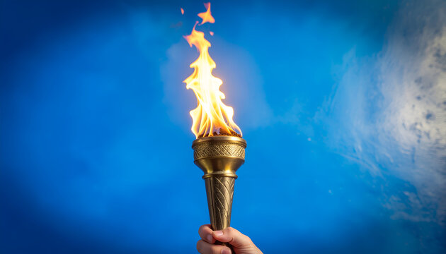 A hand holds a golden torch ablaze with vibrant flames against a striking blue sky backdrop, symbolizing hope, progress, and the spirit of competition.