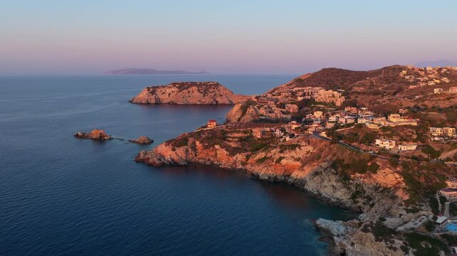 Aerial drone view of a rugged cape with a seaside village and turquoise coves in golden sunset light, Agia Pelagia, Greece