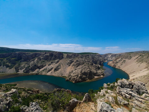 A stunning high angle view of the vibrant blue Zrmanja River canyon in Croatia, winding through steep rocky cliffs and rugged wilderness under a clear, bright summer sky.