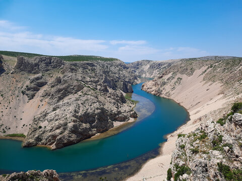 A stunning high angle view of the vibrant blue Zrmanja River canyon in Croatia, winding through steep rocky cliffs and rugged wilderness under a clear, bright summer sky.