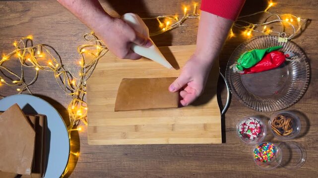Horizontal video. Top view. Man applies white icing to walls of gingerbread house on wooden cut board. Male hands with plastic bag with food paint. Cookies. Happy New Year, Merry Christmas holiday