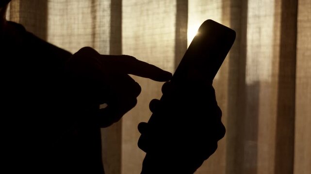 Silhouette hand scrolling on smartphone near sunlit window curtains