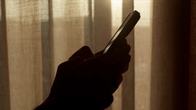 Silhouette of hand typing on smart phone near bright curtains