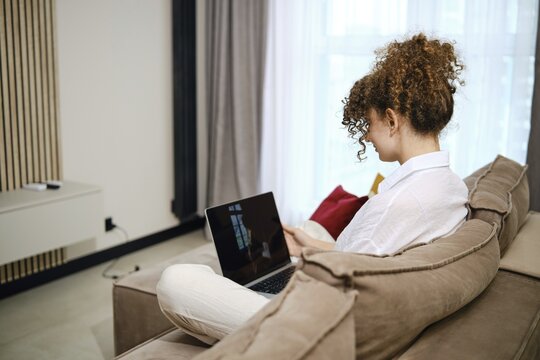 A young woman sits on the couch in the living room and reviews her photos on a laptop. The concept of pleasant memories