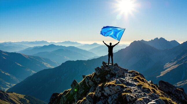 Man stands on mountain summit holding blue flag against sunlit sky. He achieved goal, reached peak, overcame challenge. Landscape shows layered mountains receding to horizon.
