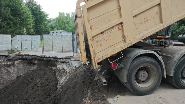 Heavy dump truck unloading dark soil into deep excavation pit during municipal water pipe repair