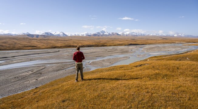Young man in front of autumnal mountain landscape, meandering Sary Jaz river, high glaciated mountain peaks of the Tien Shan in the background, autumnal mountains with yellow grass, Tien Shan, Kyrgyzstan