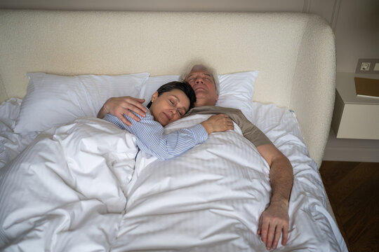 Happy relaxed senior married couple wife and husband calmly sleeping in bed and hugging each other, tranquil elderly man and woman embracing while resting in bed at night, enjoying peaceful time.