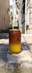  Layered iced coffee with fresh orange juice in a glass on a café table in a narrow Mediterranean old town street. Refreshing summer espresso drink with ice and paper straw. Vertical clo © Elena