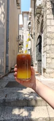 Hand holding a layered iced espresso with fresh orange juice in a historic Mediterranean street. Refreshing cold summer coffee drink with ice and paper straw. Vertical lifestyle travel  © Elena
