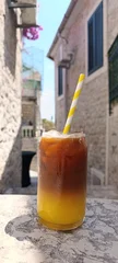 Refreshing espresso and orange juice served over ice on a stone table in a historic Mediterranean alley. Layered summer coffee drink with paper straw. Vertical travel lifestyle p © Elena