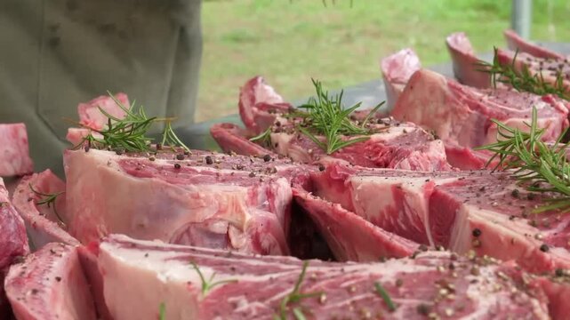 A close-up shot of several premium, thick-cut raw beef steaks, including T-bone and ribeye, heavily seasoned with cracked black pepper and fresh rosemary sprigs, ready for grilling at an outdoor barbe