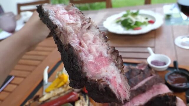 A mouthwatering, close-up shot of a perfectly grilled and sliced medium-rare tomahawk ribeye steak served on a rustic platter. The feast includes charred tiger prawns, roasted yellow peppers, and dipp