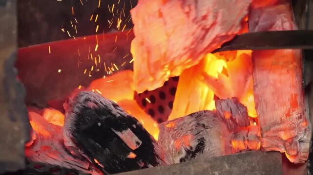High-angle close-up shot of burning charcoal briquettes igniting with orange flames and rising smoke. The glowing embers showcase the initial stages of heat preparation for grilling steaks at a summer