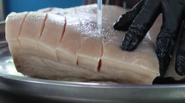 A detailed close-up shot of a large slab of fresh raw pork belly with deep scoring on the skin, resting on a stainless steel tray. A person wearing black nitrile gloves is shown handling the meat, hig
