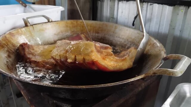 A close-up, high-angle shot of large slabs of marinated pork belly deep-frying in hot, shimmering oil within a seasoned metal wok or pan. A cook uses a large spoon to baste and move the meat, showing 