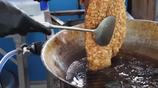 A close-up footage of a large slab of seasoned pork belly being deep-fried in a large wok. The skin is golden-brown and bubbling into a perfectly crispy crackling texture, a staple of Thai and Filipin