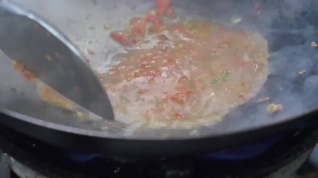 A professional close-up shot of a chef using a spatula to stir-fry a vibrant mix of minced red chilies, garlic, and shallots in a hot metal wok. Steam and sizzling oil highlight the intense heat and a